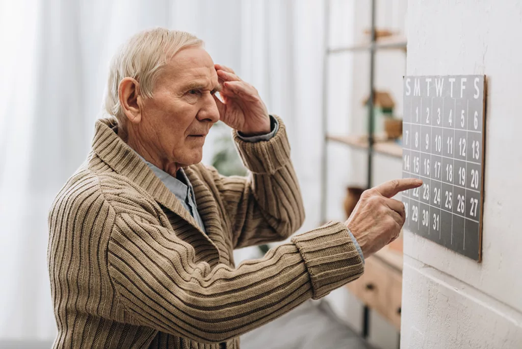 Older man holding hand to head and other pointing to wall calendar. Looks confused.